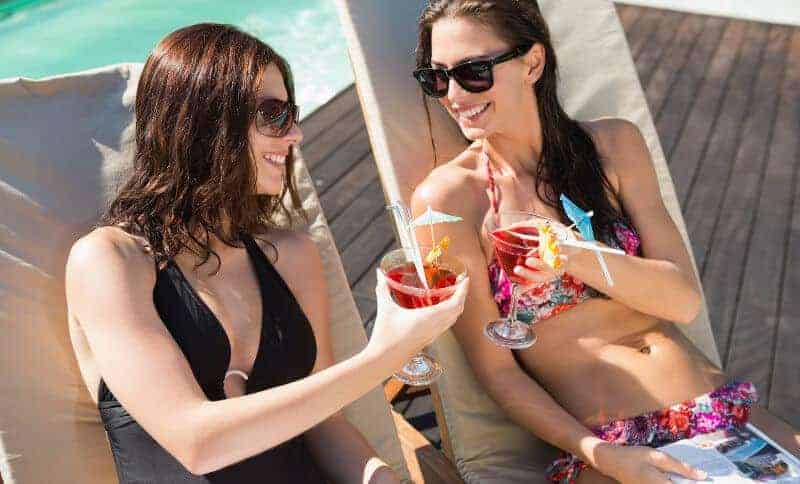 Two young women sitting poolside on loungers, making a toast with fruity drinks