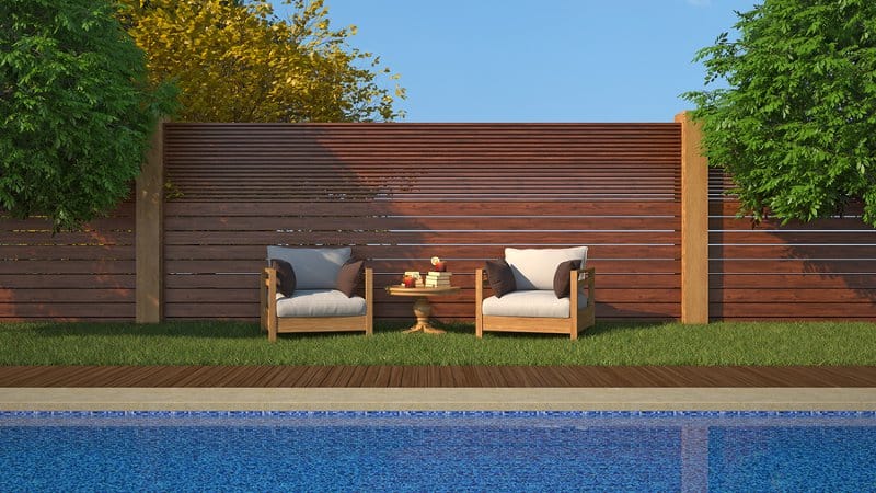 Two luxury outdoor chairs near an inground swimming pool with a high-end wooden pool fence in the background