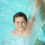 Boy in a swimming pool smiling under a waterfall
