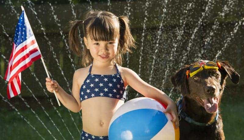 Young girl holds a flag and a beach ball, standing next to a dog wearing glasses with water spraying in the background