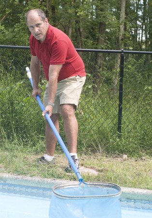 Middle aged man in a red shirt skimming leaves out of an inground pool