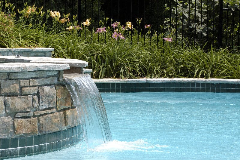 Closeup of a waterfall built into an inground swimming pool, with flowers and a wrought iron fence in the background