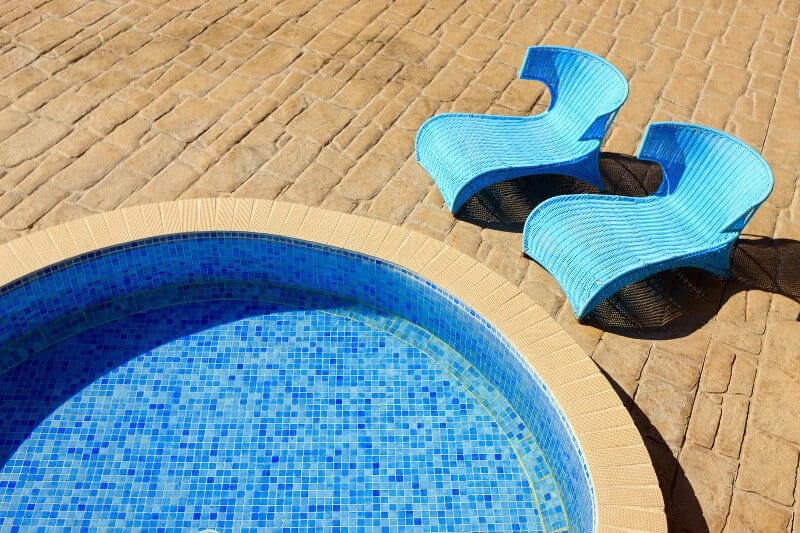 Small, round inground swimming pool with two blue loungers sitting nearby on the stone pool deck