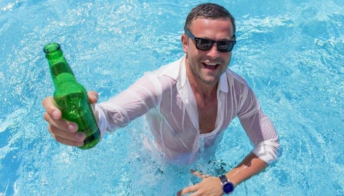 Fully dressed man raising a beer bottle in a swimming pool