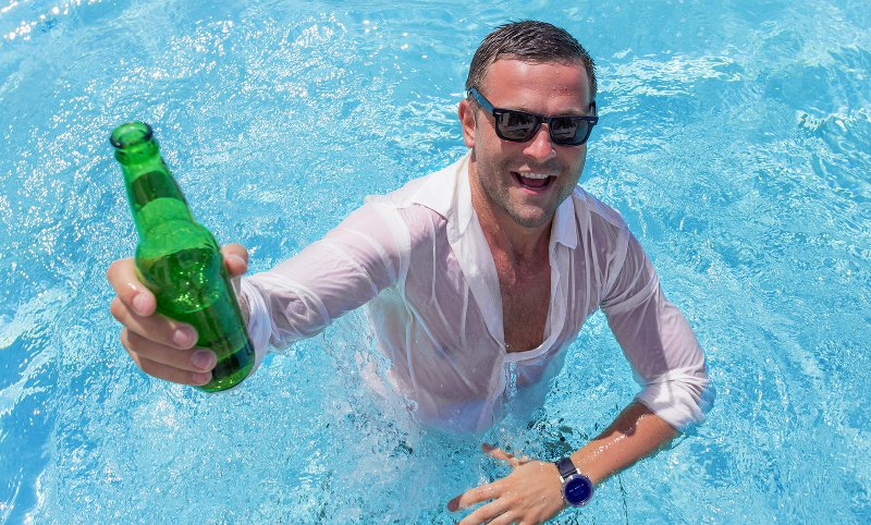 Fully dressed man raising a beer bottle in a swimming pool