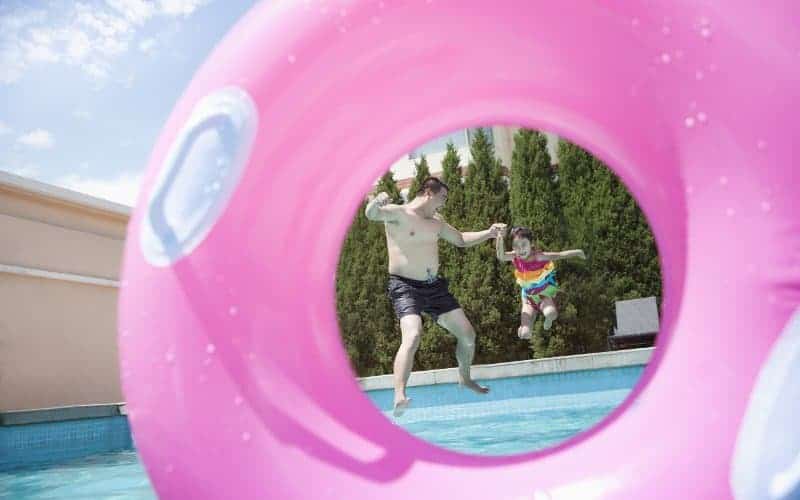 Father and young daughter jumping into an inground swimming pool, as seen through a pink inflatable tube